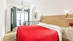 Une chambre avec une couvre-lit rouge, une tête de lit en bois, une table de chevet blanche ornée d’un vase avec une fleur rouge, et une vue sur un balcon.