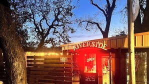 Un edificio con un letrero que dice 'RIVERSIDE' y un rótulo rojo que indica 'ABIERTO, ESPERE A SER ASIGNADO'.