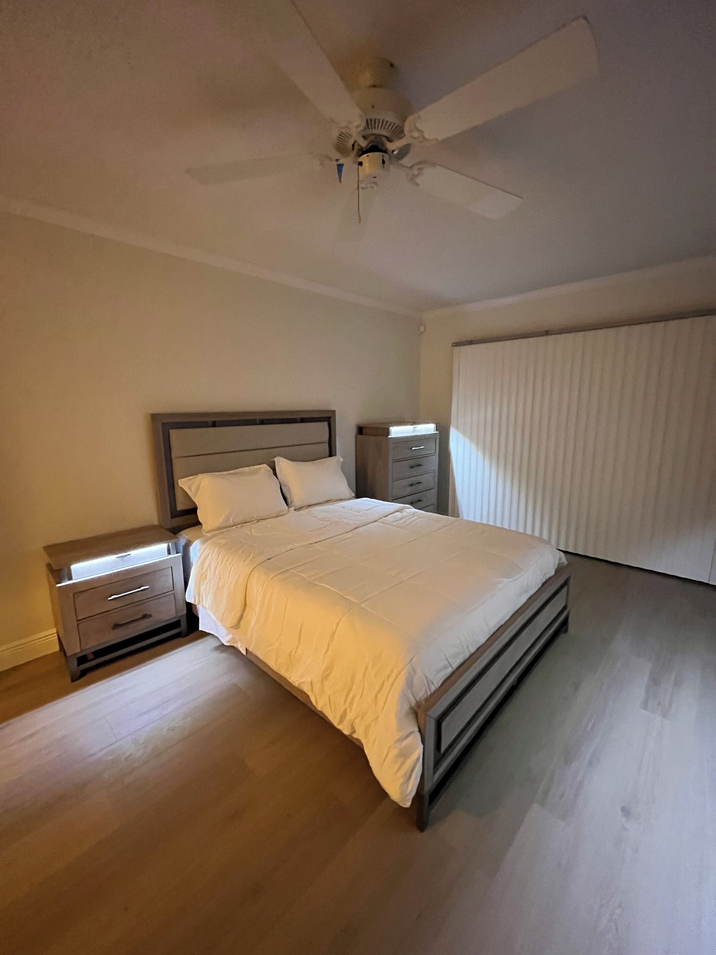 Een slaapkamer met een bed, nachtkastjes, een plafondventilator en een raam met luxaflex.