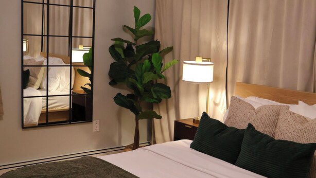 En pænt redt seng med et grønt plaide, et stort spejl, en potteplante og en sengetårnslampe på et hotelværelse.