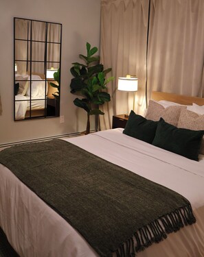 Un lit bien fait, agrémenté d’un plaid vert, un grand miroir, une plante en pot et une lampe de chevet, dans une chambre d’hôtel.