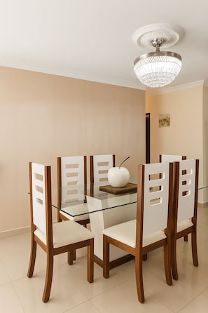 A dining area with a glass table surrounded by wooden chairs with cut-out backrests. A decorative vase is placed on the table, and a chandelier hangs from the ceiling.