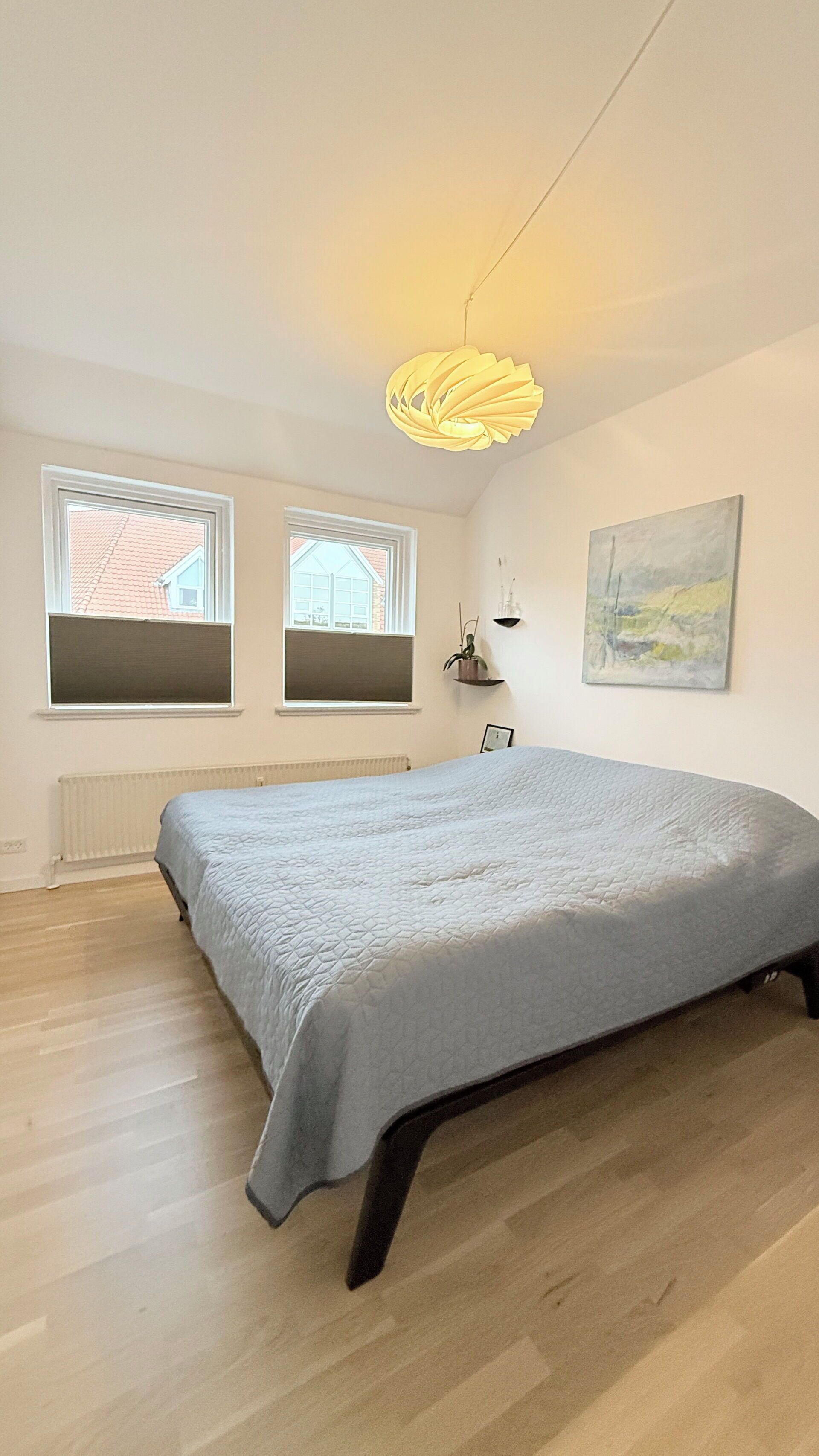 A bedroom with a bed, two windows, a radiator, a hanging light fixture, and a painting on the wall.