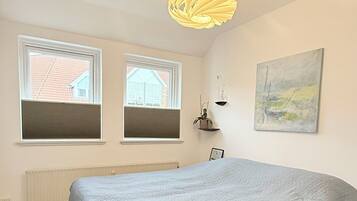 A bedroom with a bed, two windows, a radiator, a hanging light fixture, and a painting on the wall.