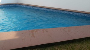 Una piscina con agua cristalina azul, rodeada por un suelo de baldosas y una zona de césped.