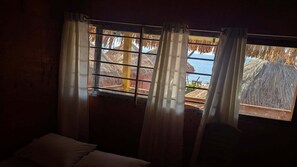 A room with a large window, curtains, and a view of a beach with thatched roofs. - Beach Valley Eco Resort Saint Martin (Saint Martin's Island)