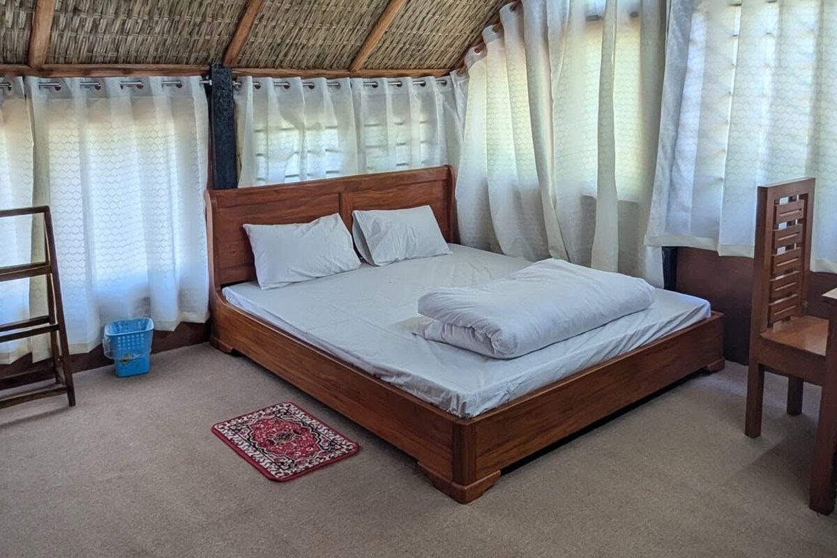 Una cama individual con cabecera de madera en una habitación con techo de paja y ventilador de techo.