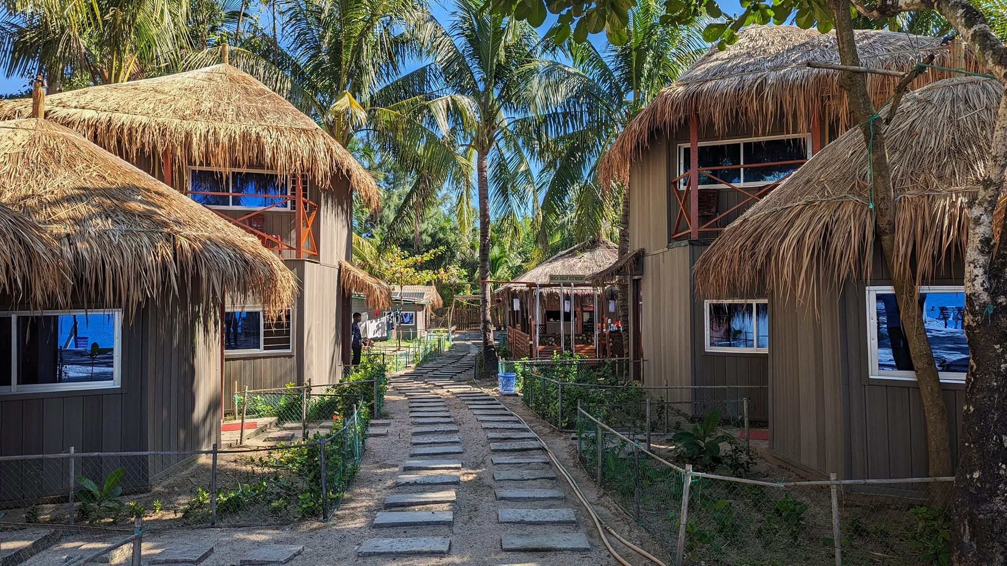 A pathway leading to thatched-roof huts with windows, surrounded by palm trees.