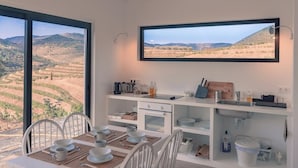 Uma área de refeições com uma mesa posta para quatro pessoas, uma cozinha com pia e bancada, e uma janela grande com vista para campos em socalcos.
