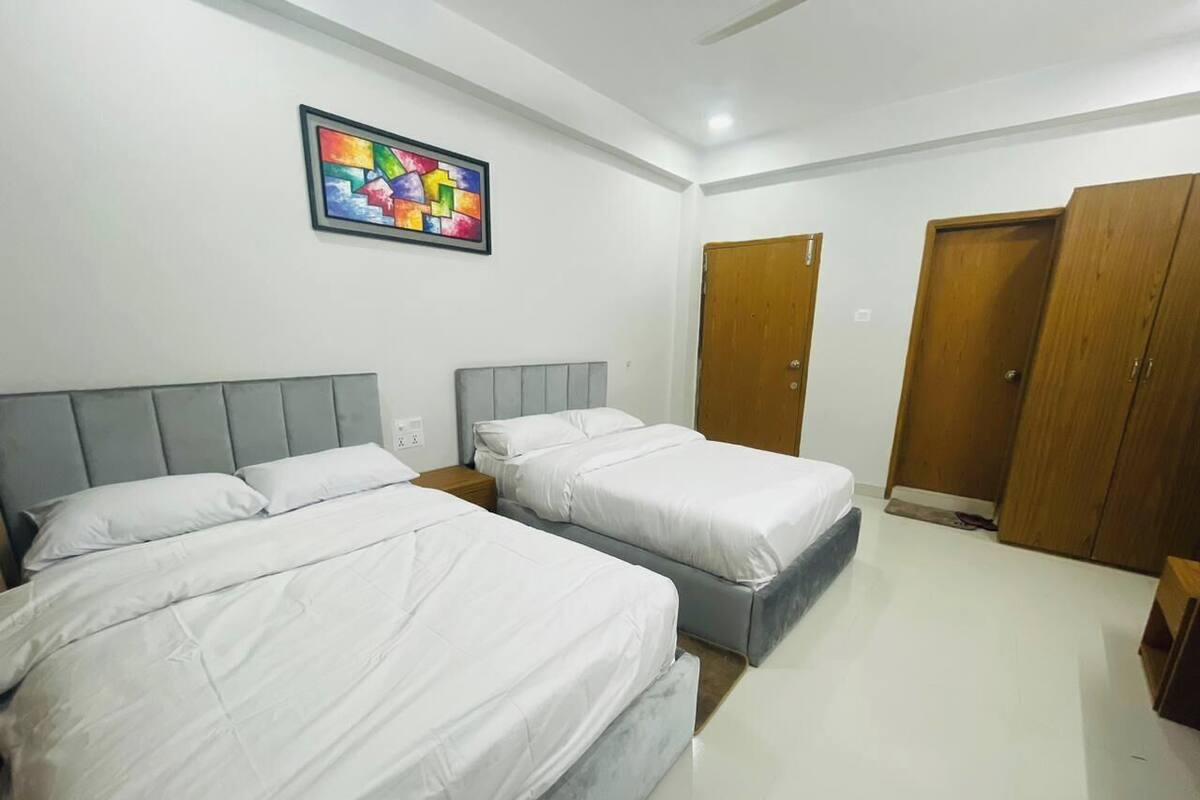 A hotel room with two beds, a wooden wardrobe, a colorful abstract painting, and a white tiled floor.