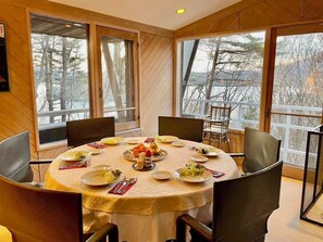 A dining area with a round table set for six, surrounded by large windows offering a view of trees and a balcony.