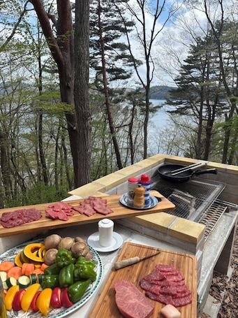 An outdoor cooking area with a grill, various meats, vegetables, and condiments.