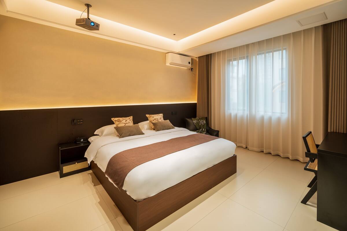 Una habitación de hotel moderna con una cama grande, mesitas de noche, un escritorio y un proyector.