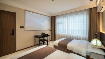 A hotel room with two beds, a desk, a projector screen, and a window with curtains.