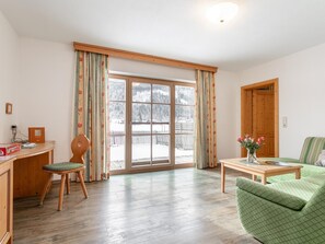 A room with a wooden desk, a chair, a green sofa, a wooden coffee table, and a large window with curtains. - Holiday Home in Salzburg with Alpine View (Leogang)