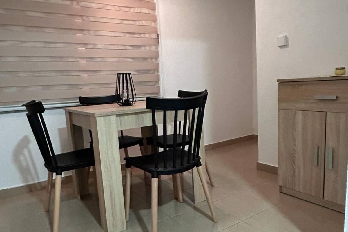 A small dining area with a wooden table and four black chairs. There is a cabinet to the right and a window with blinds in the background.