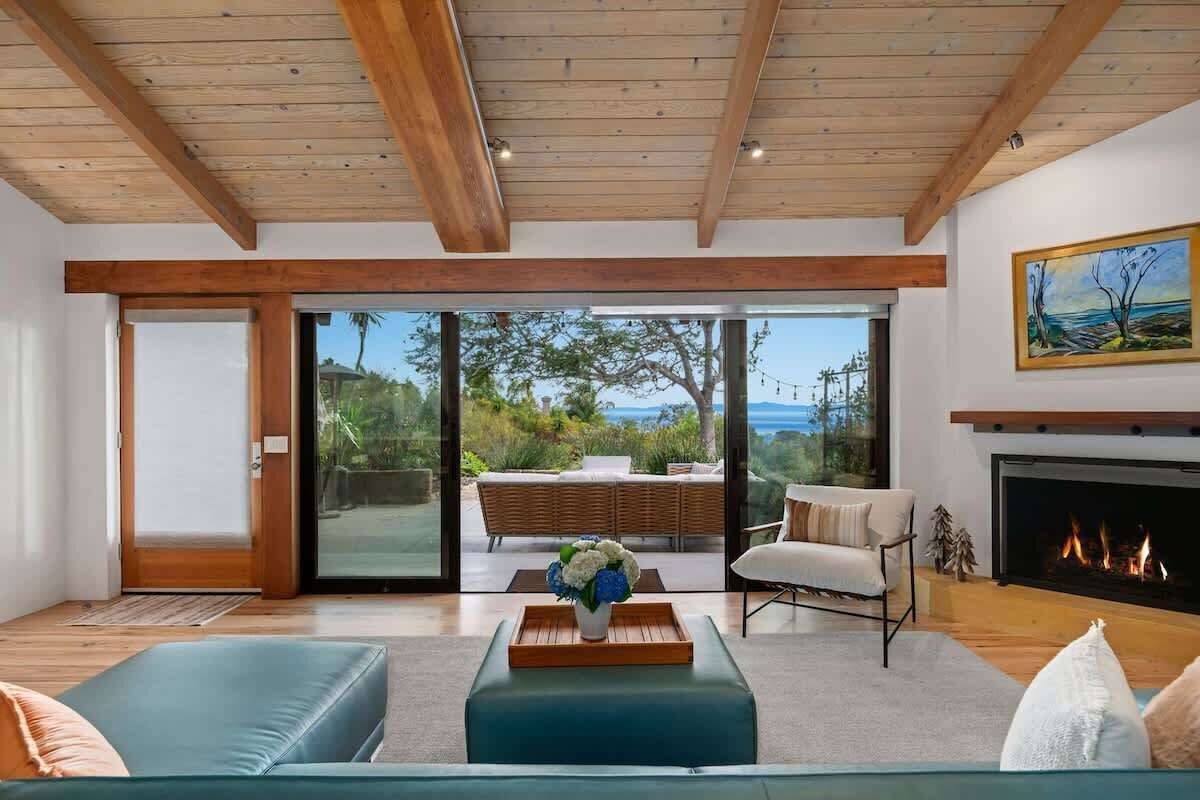 A modern living room with a fireplace, a sofa, a coffee table with flowers, and a large window offering a view of the outdoors.