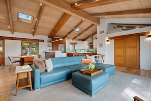 A spacious living area with a blue sectional sofa, wooden beams on the ceiling, and a view of the kitchen in the background.