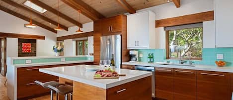 A modern kitchen with wooden cabinets, a central island, bar stools, and a large window providing natural light.
