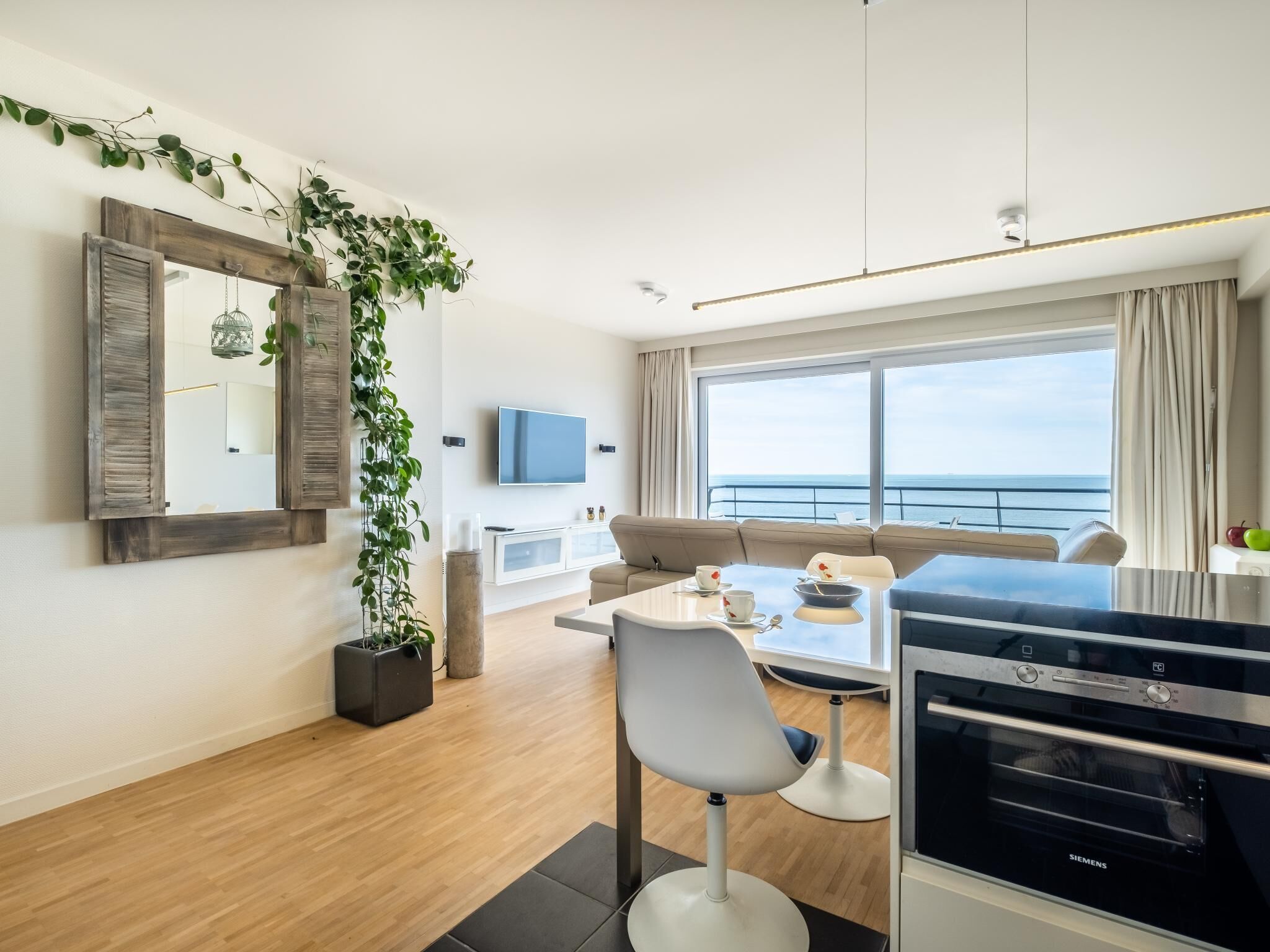 Una sala moderna con un sofá, una mesa de comedor con sillas, un televisor, un ventanal con vista al mar y una zona de cocina con horno Siemens.