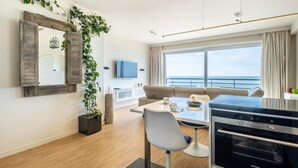 Una sala moderna con un sofá, una mesa de comedor con sillas, un televisor, un ventanal con vista al mar y una zona de cocina con horno Siemens.