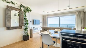 Una sala moderna con un sofá, una mesa de comedor con sillas, un televisor, un ventanal con vista al mar y una zona de cocina con horno Siemens.
