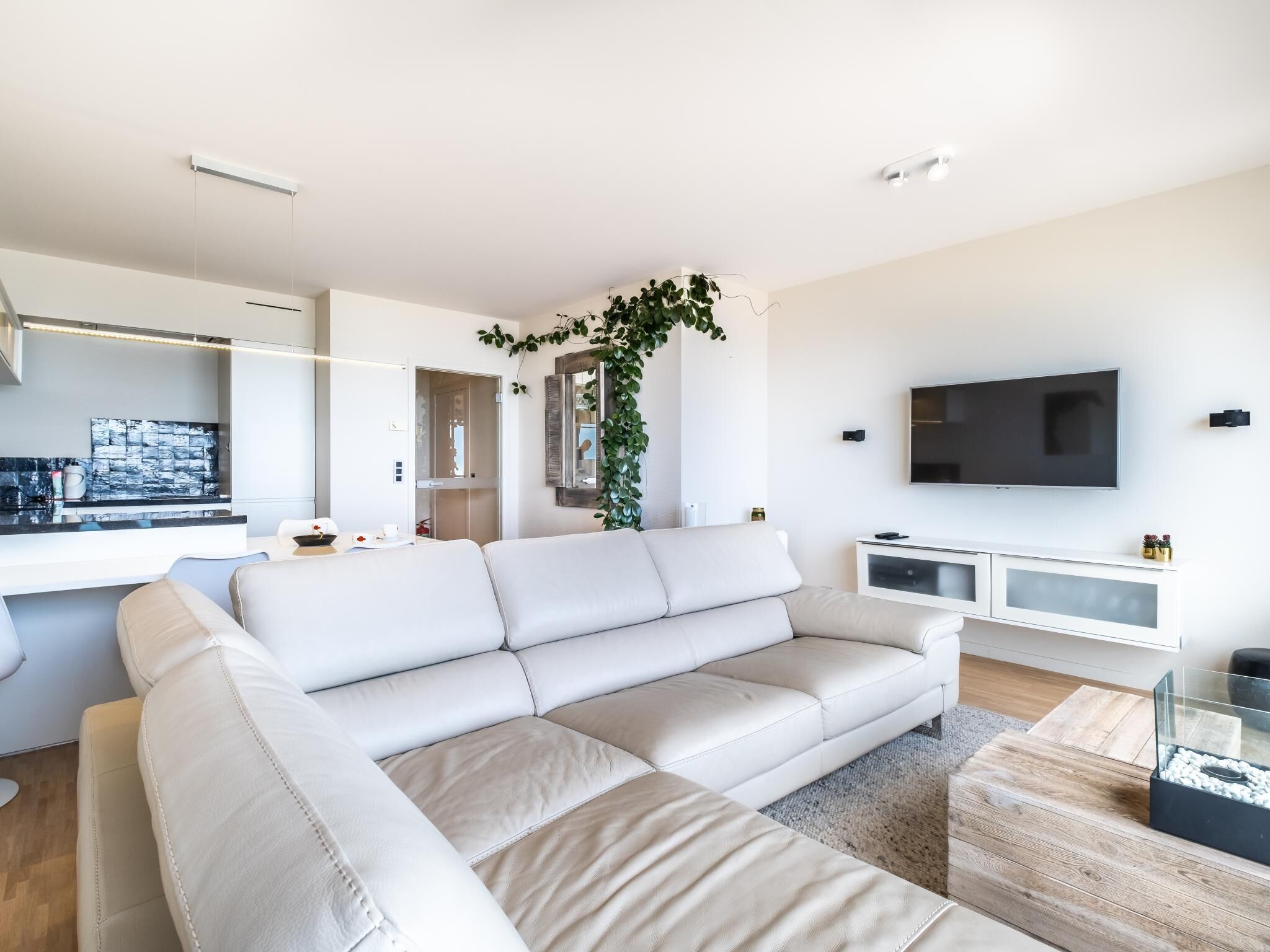 Una sala de estar moderna con un sofá blanco, un televisor y una cocina al fondo.