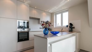 A modern kitchen with white cabinetry, a built-in oven, a coffee maker, and a blue vase with dried flowers on the island. - Cosy flat with beautiful terrace (Koksijde)