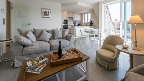 A modern living room with a grey sofa, a wooden coffee table with magazines, a wine bottle, and glasses. There is a dining area with a white table and chairs, and a kitchen in the background. - Cosy flat with beautiful terrace (Koksijde)