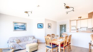 A well-lit living room with a sofa, ottoman, and dining table set with chairs. There are framed pictures on the wall, a kitchen area with cabinets, and a window. - Wake up to sea views (Koksijde)