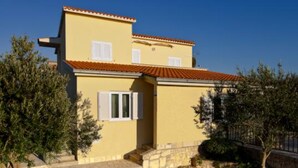 A two-story building with a yellow facade and red-tiled roof. There are two windows with shutters, and a small staircase leading to the entrance. - Apartman Olive Garden (Solta)