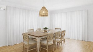 A dining area with a wooden table and wicker chairs, white curtains, and a hanging light fixture.