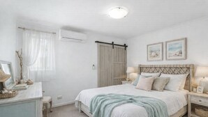 A bedroom with a bed, bedside tables, a dresser, a window with sheer curtains, an air conditioning unit, and framed artwork on the wall.