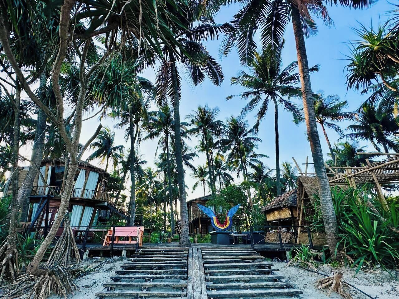 Ein Resort mit mehreren Gebäuden, Palmen und einer Treppe zu einem Strandabschnitt.