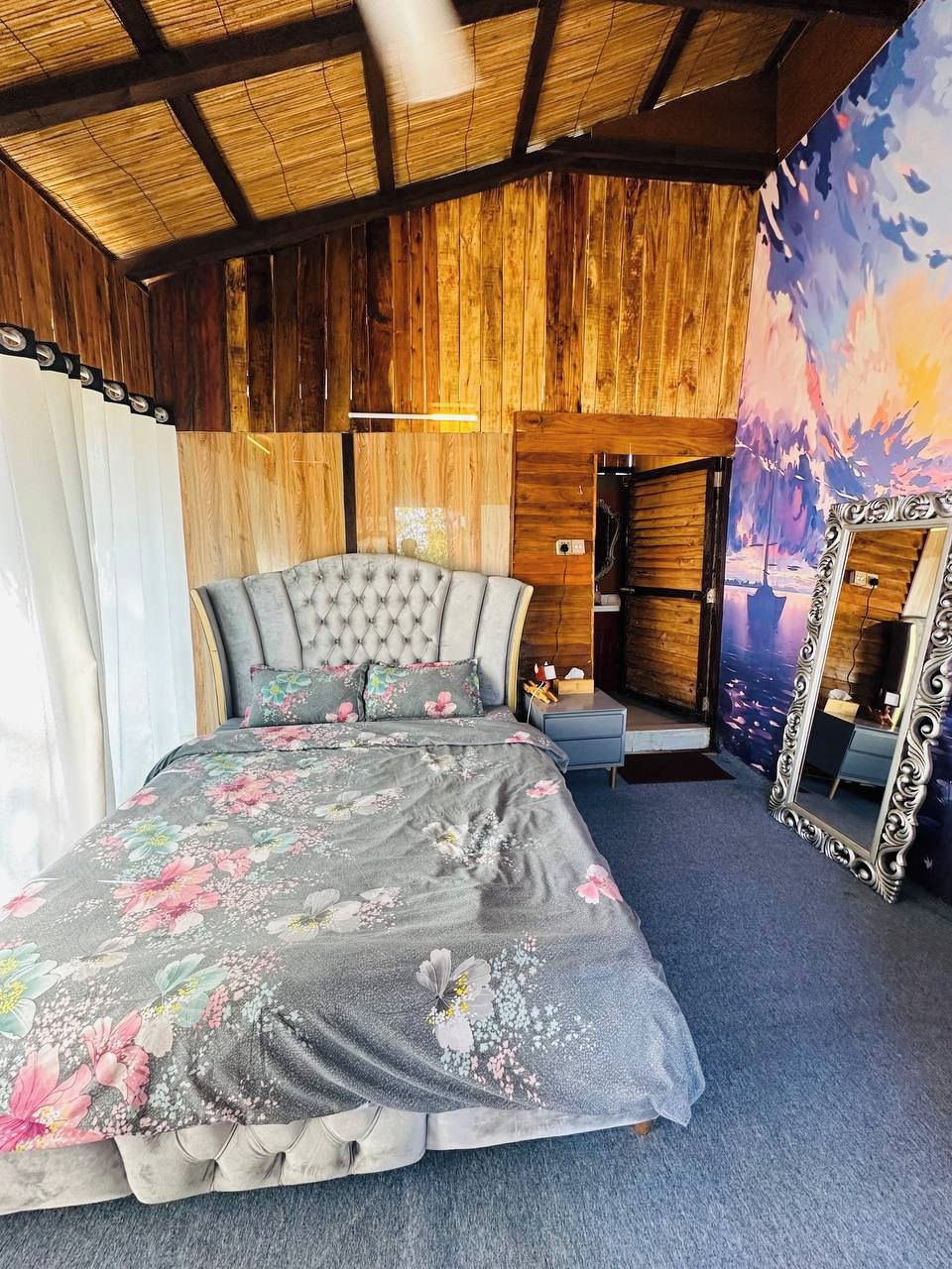 A bedroom with a tufted headboard, floral bedding, a large mirror, and a wooden ceiling.