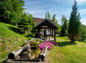 A wooden gazebo with a tiled roof, surrounded by a garden with flowers and logs. - Holiday House Briški (Delnice)