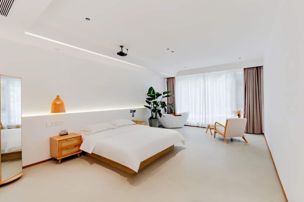 Habitación de hotel moderna con una cama grande, una mesita de noche, una silla, una planta en maceta y un ventanal con cortinas.