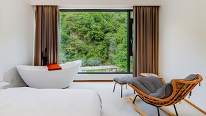 A modern room with a large window, a white bathtub, a wicker chair, and a bed.
