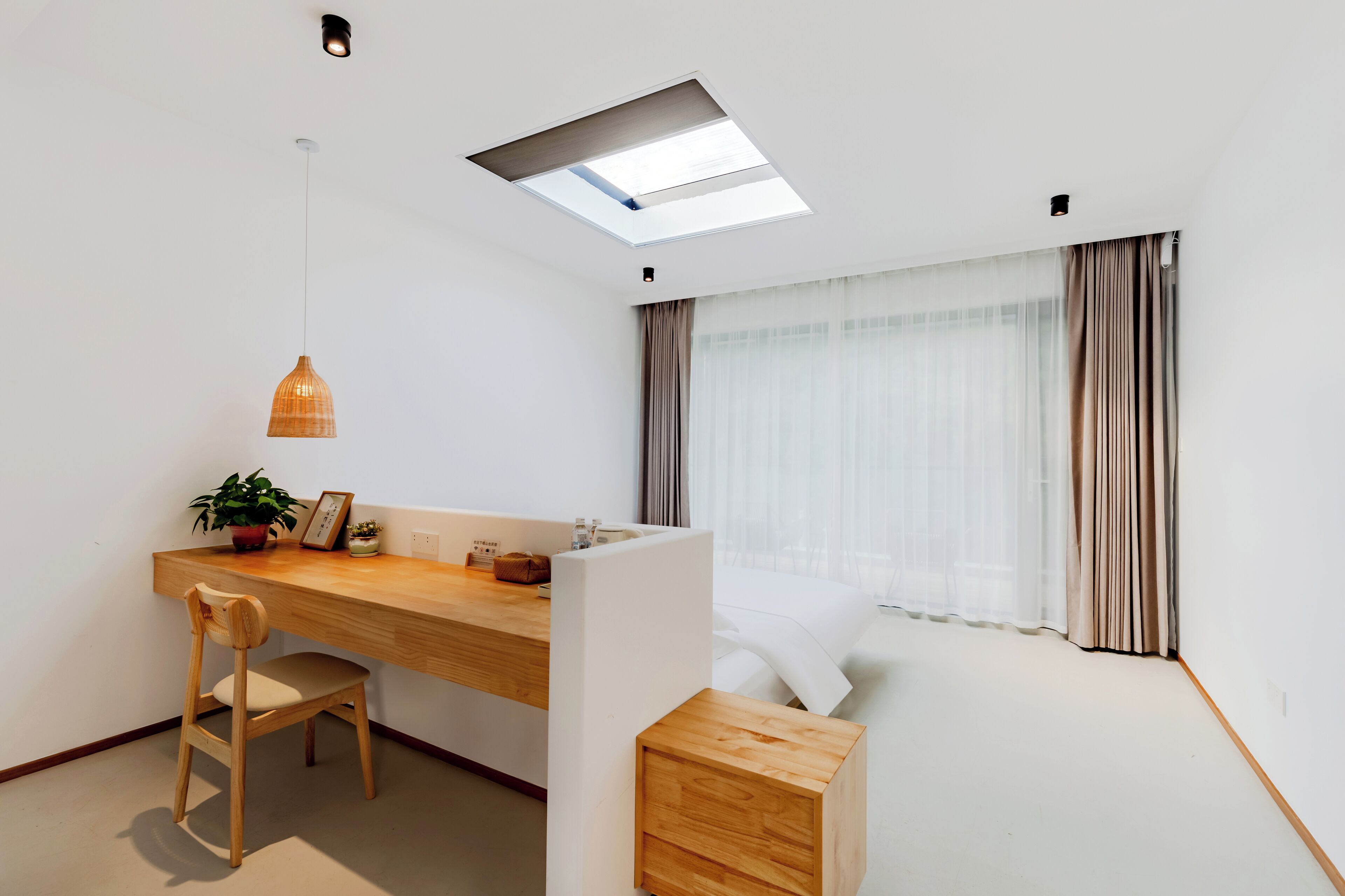Habitación moderna y minimalista con un escritorio de madera, una silla, un taburete pequeño de madera, una planta en maceta y una cama con cortinas ligeras.