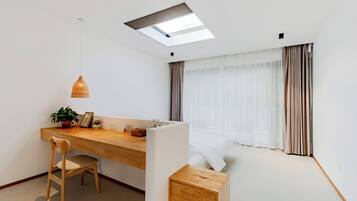 Habitación moderna y minimalista con un escritorio de madera, una silla, un taburete pequeño de madera, una planta en maceta y una cama con cortinas ligeras.