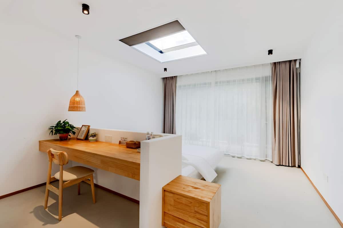 Un dormitorio moderno y minimalista con un escritorio de madera, una silla, una mesita de noche y un amplio ventanal con cortinas.