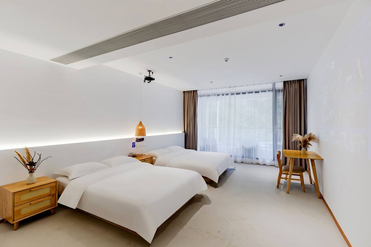 Una habitación de hotel moderna con dos camas, una mesita de noche de madera, una pequeña mesa con una silla y un ventanal con cortinas.