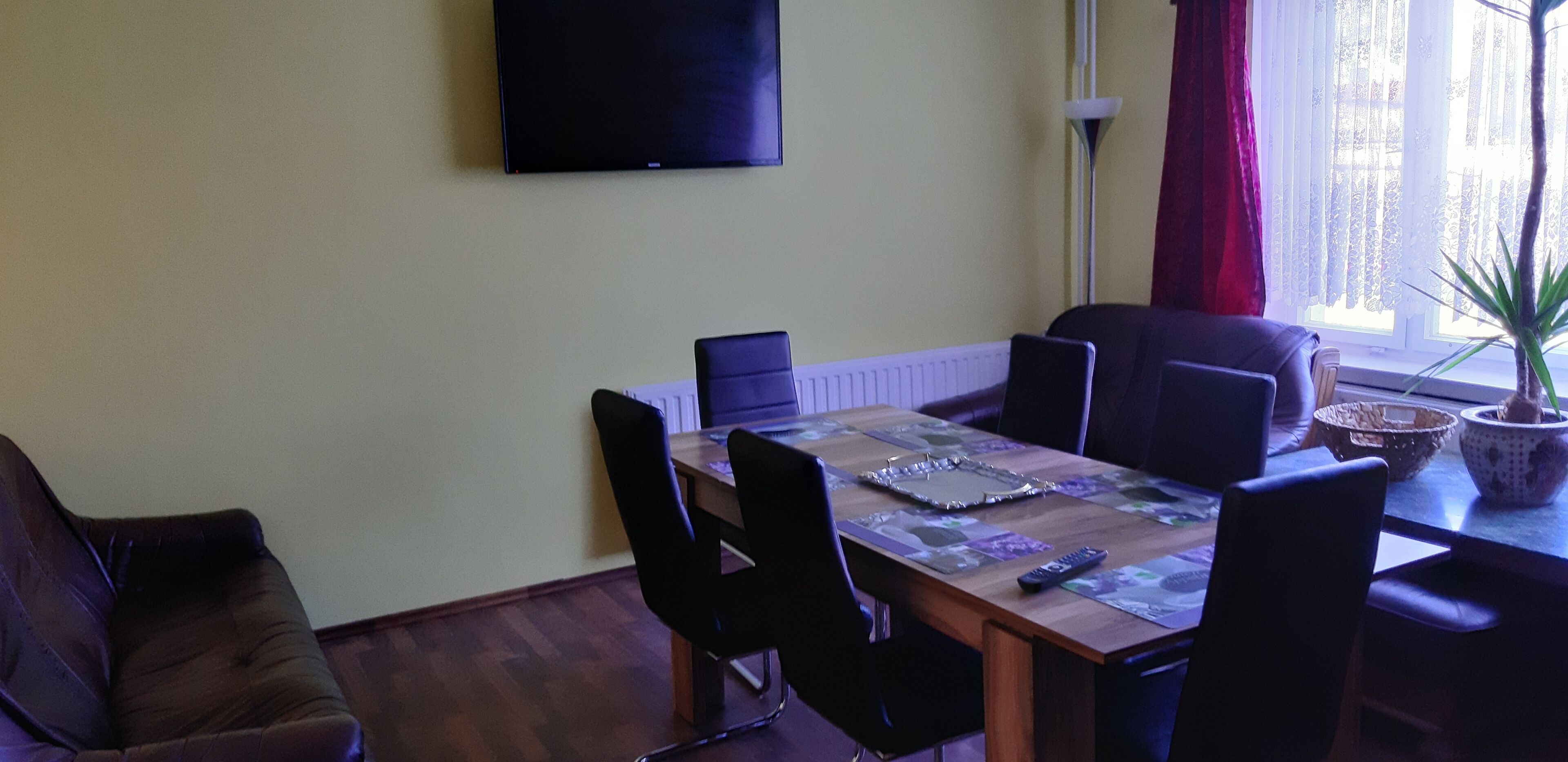 A dining area with a table set for four, surrounded by chairs, a flat-screen TV mounted on the wall, and a window with curtains.