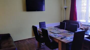 A dining area with a table set for four, surrounded by chairs, a flat-screen TV mounted on the wall, and a window with curtains.