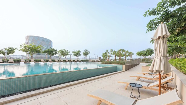 Ein Poolbereich mit Liegestühlen und Sonnenschirmen, ein gefliestes Schwimmbecken und ein modernes Gebäude im Hintergrund.