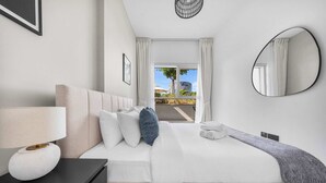 Quarto de hotel moderno com uma cama grande, mesa de cabeceira com luminária, espelho e vista para o exterior.