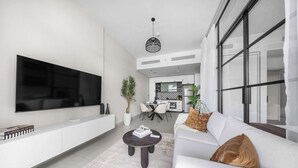 Ein modernes Wohnzimmer mit großem Flachbildfernseher, einem dunklen Couchtisch, weißen Sofas und einem Essbereich im Hintergrund.
