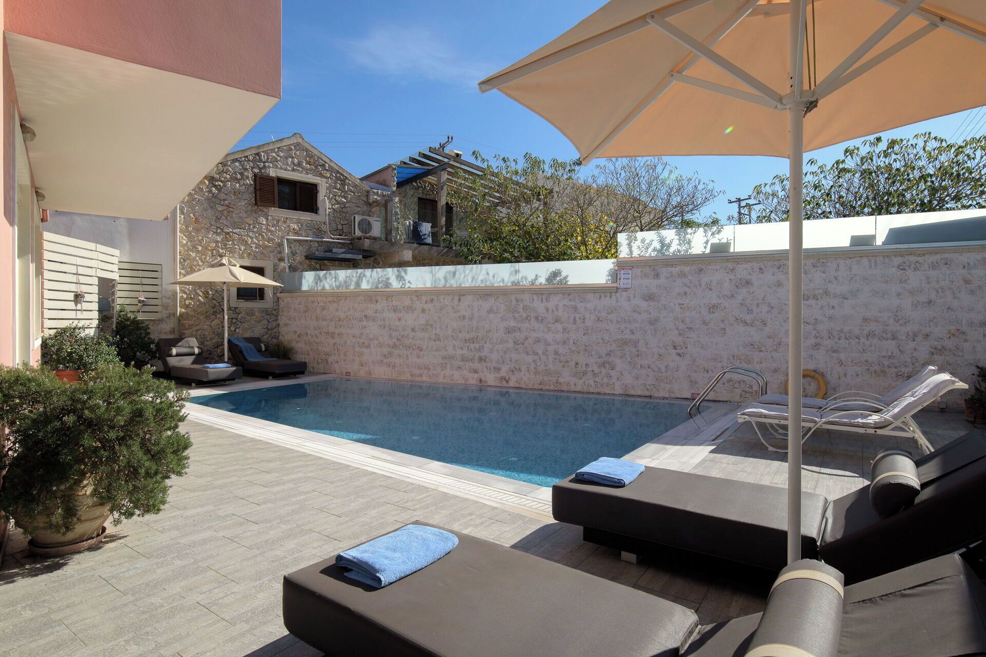 Piscina com espreguiçadeiras e guarda-sóis, cercada por muros de pedra e plantas em vasos.