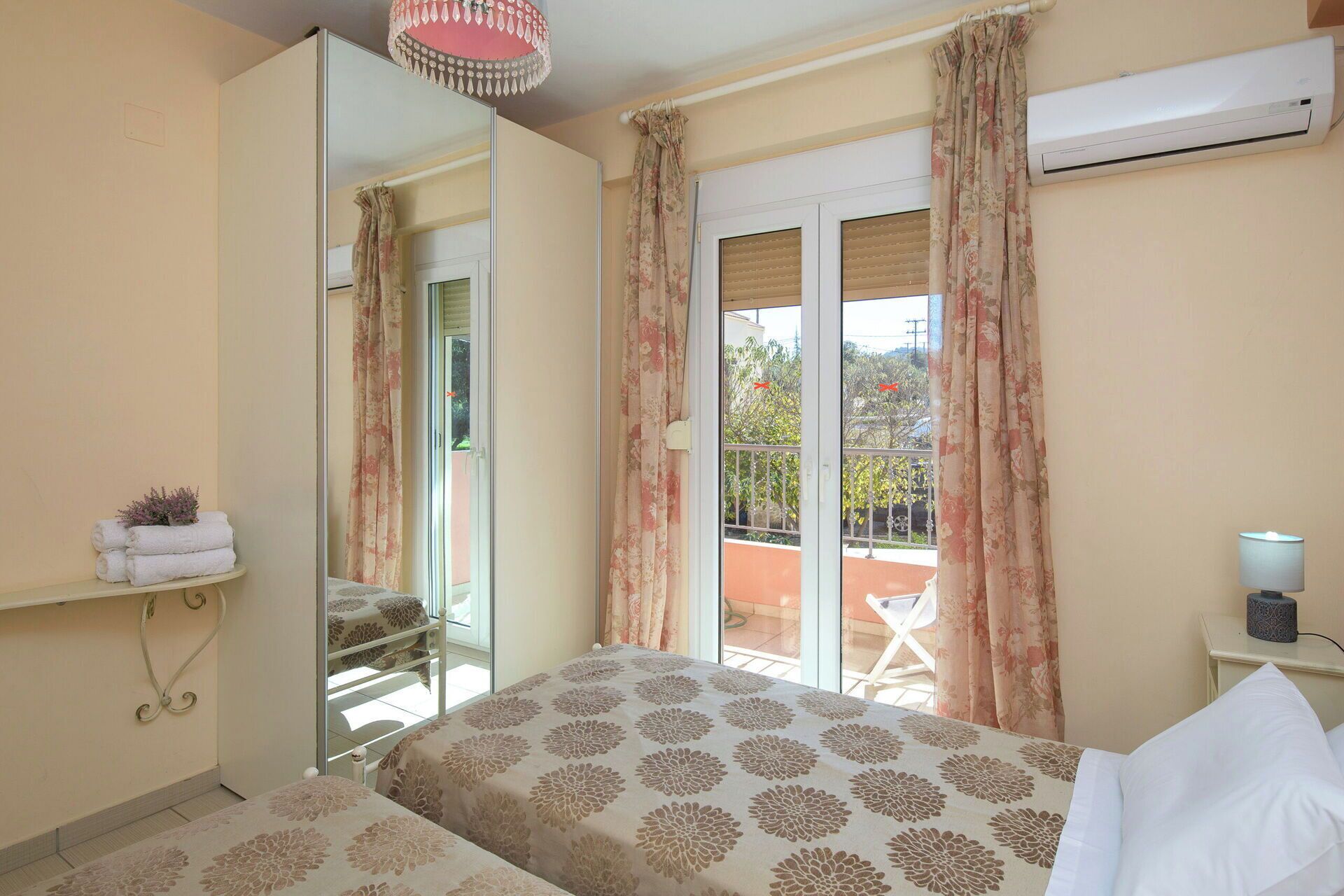 A bedroom with a bed, a mirror, a balcony with curtains, an air conditioning unit, and a small table with a lamp.