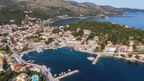 Vista aérea de un pueblo costero con un puerto deportivo, rodeado de colinas y aguas azules cristalinas.